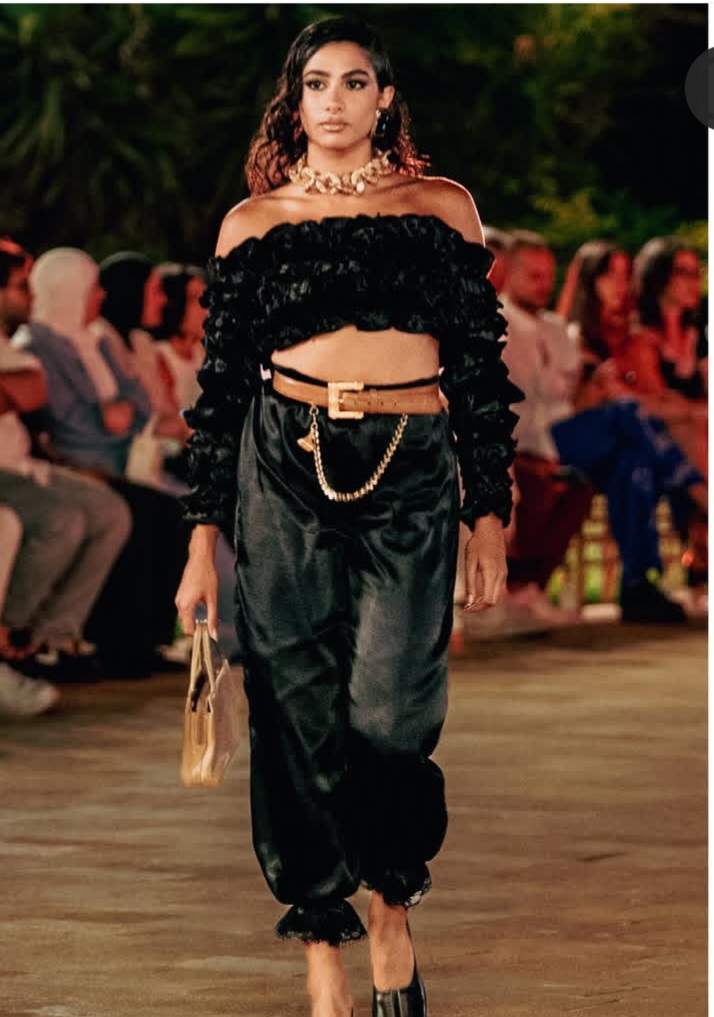 Une silhouette noire incarne cette fusion de puissance et de décontraction : un haut noir brillant orné de volants volumineux est associé à un pantalon en satin drapé noir. Les accessoires dorés – un collier chaîne épais et une boucle de ceinture imposante – ancrent le look avec une autorité critique. 